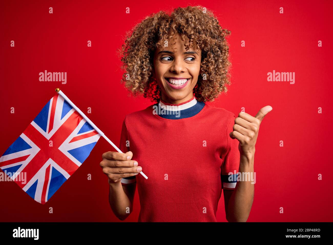 Giovane donna riccia afroamericana che detiene la bandiera britannica per celebrare il referendum brexit indicando e mostrando con il pollice fino alla parte con felice faccia smilin Foto Stock
