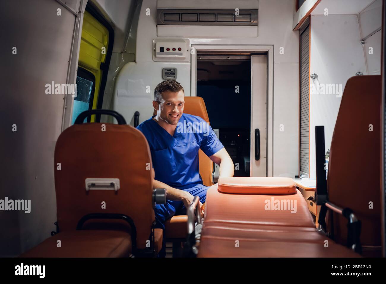 Uomo in un uniforme medica seduto in auto ambulanza e sorridendo Foto Stock