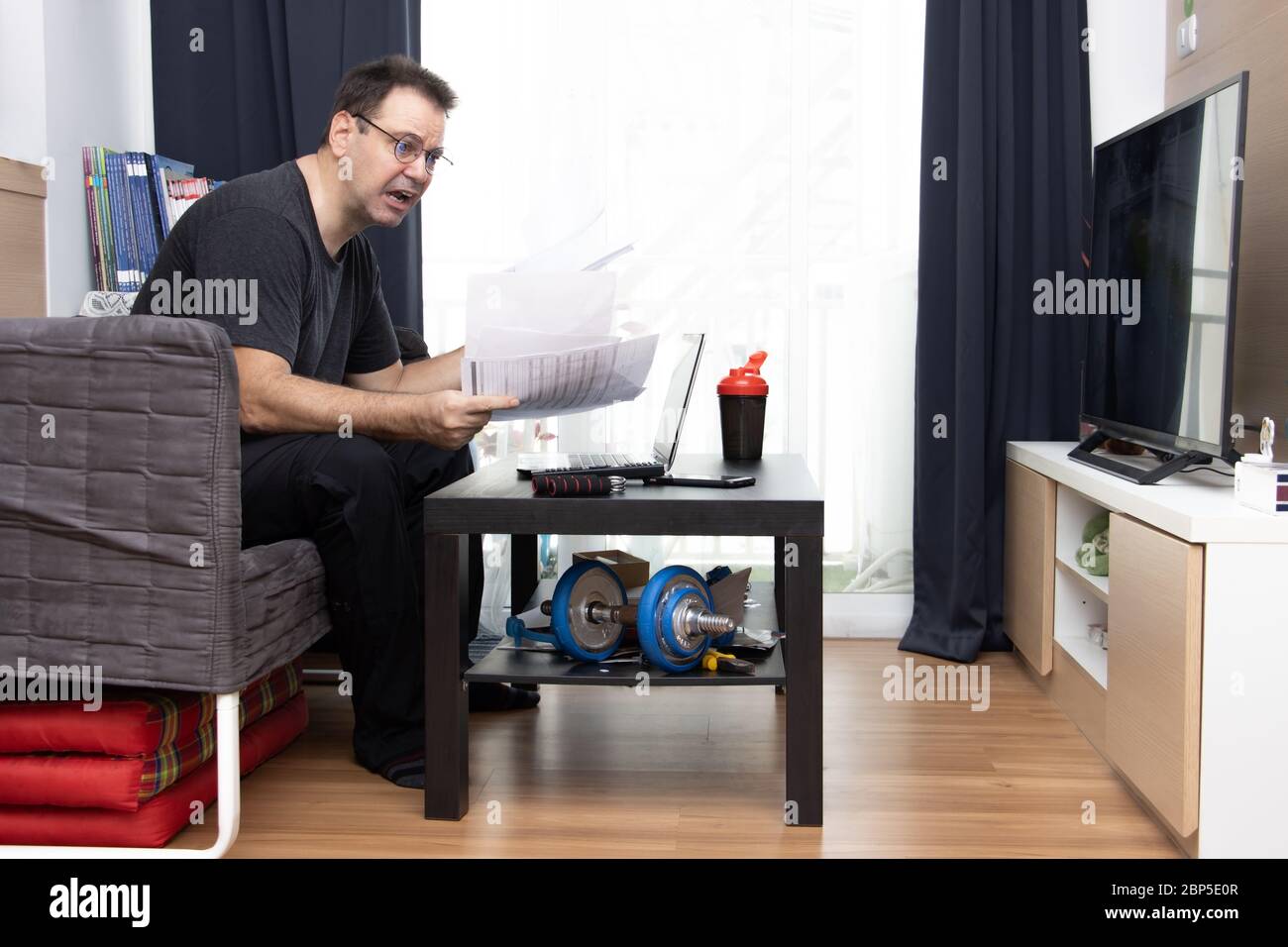 Un uomo stressato seduto in soggiorno e lavora con documenti cartacei e laptop su un tavolo. Ufficio domestico. Foto Stock
