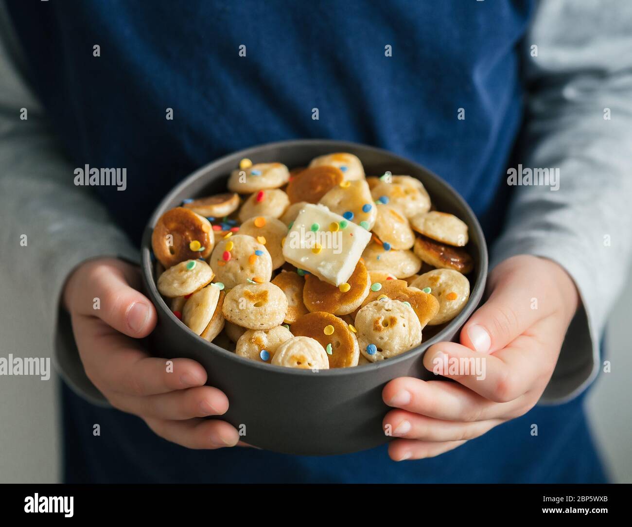 Cibo alla moda - cereali pancake. Un mucchio di mini frittelle di cereali in boul nelle mani dei bambini. Irriconoscibile ciotola grigia con minuscoli pancake decorati con spruzzette e burro. Spazio di copia per testo o disegno Foto Stock