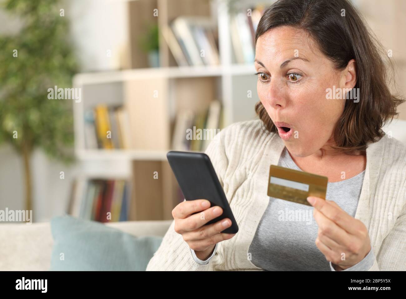 Donna adulta sorpresa che paga online su smartphone con carta di credito seduta sul divano a casa Foto Stock