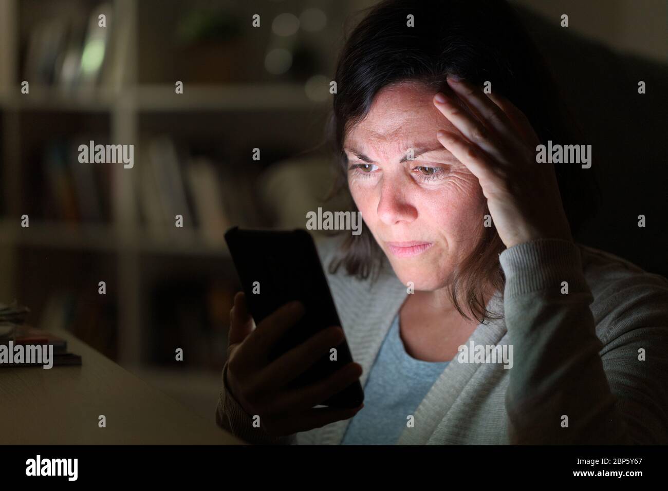 Donna adulta preoccupata che legge sullo smartphone schermo illuminato seduto di notte a casa Foto Stock