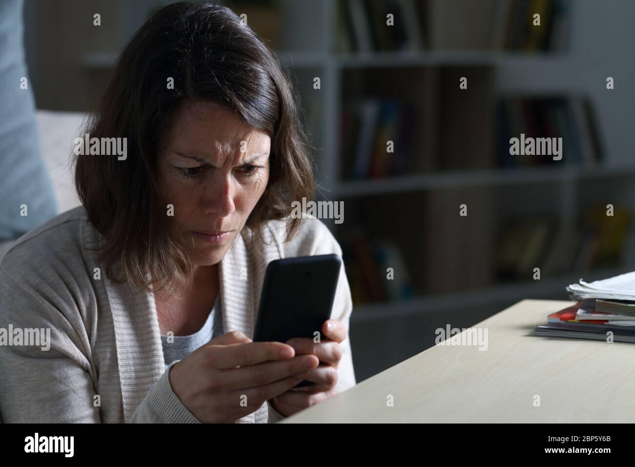 Donna adulta preoccupata che legge le cattive notizie sullo smartphone seduto sul pavimento di notte a casa Foto Stock