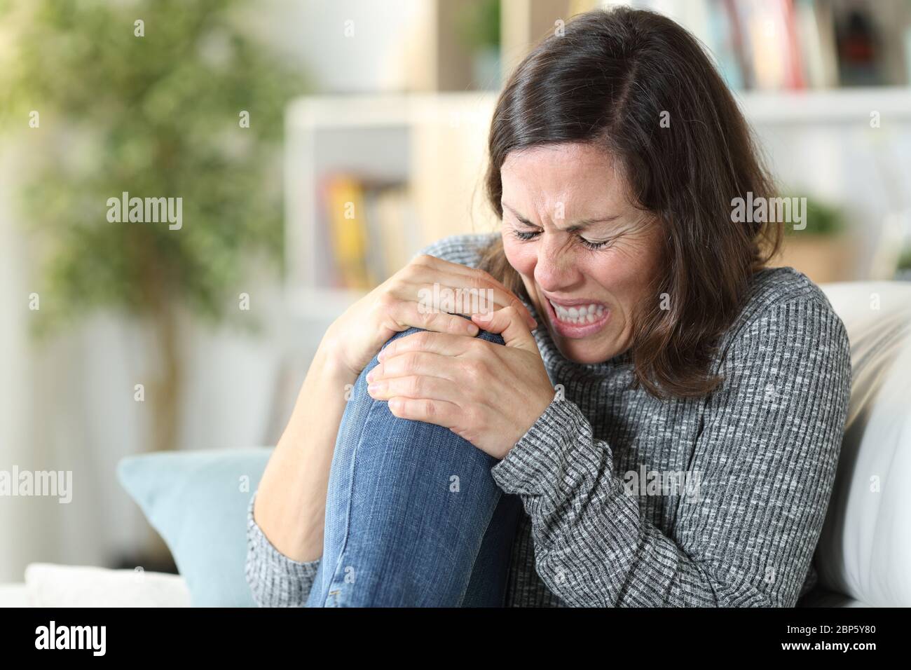 Donna adulta in dolore che soffre di dolore al ginocchio seduto su un divano a casa Foto Stock