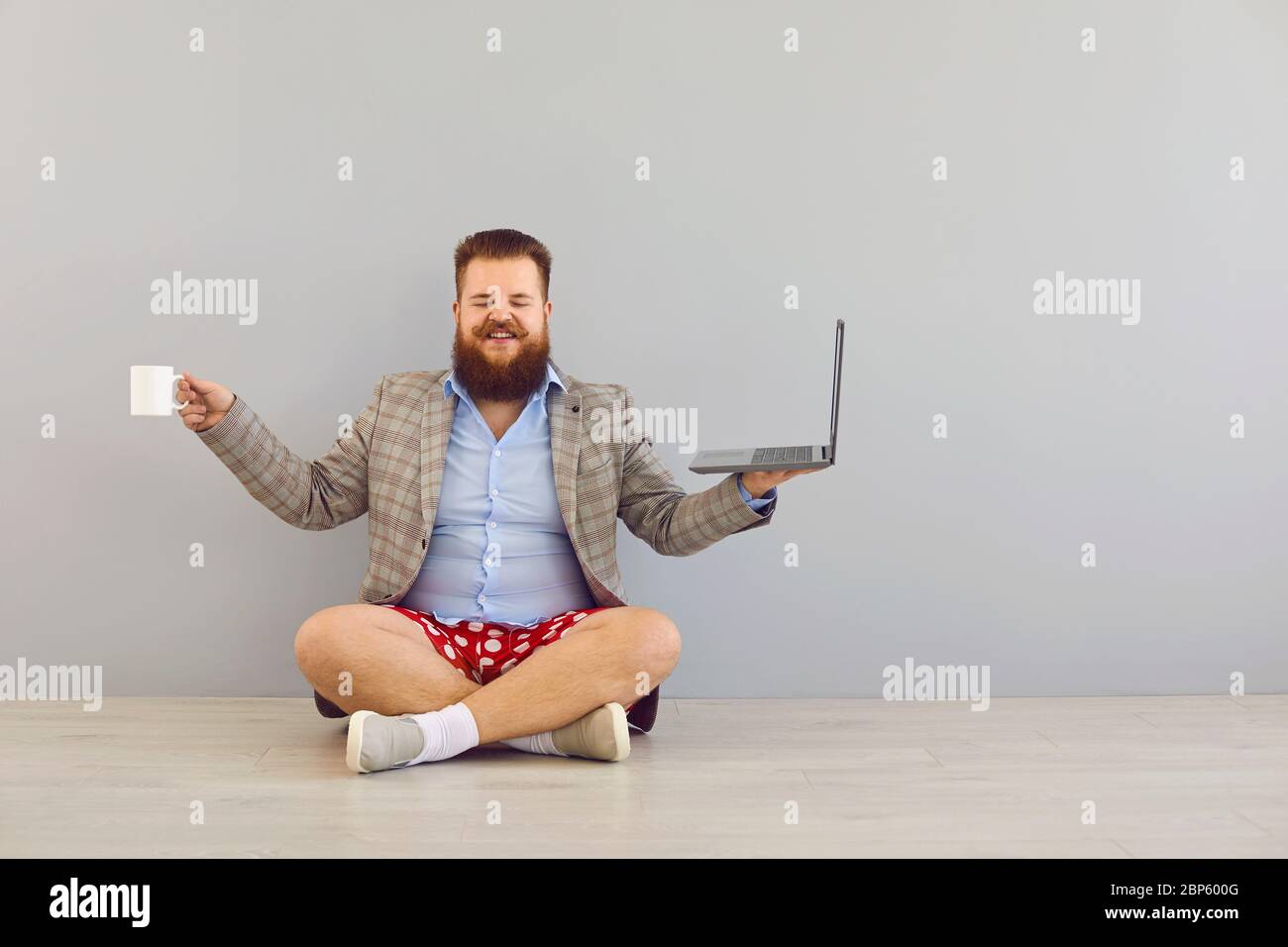 Concetto di lavoro a distanza freelance online. Divertente uomo con cuscinetti in tessuto adiposo in giacca e shorts rossi con un computer portatile su sfondo grigio. Foto Stock