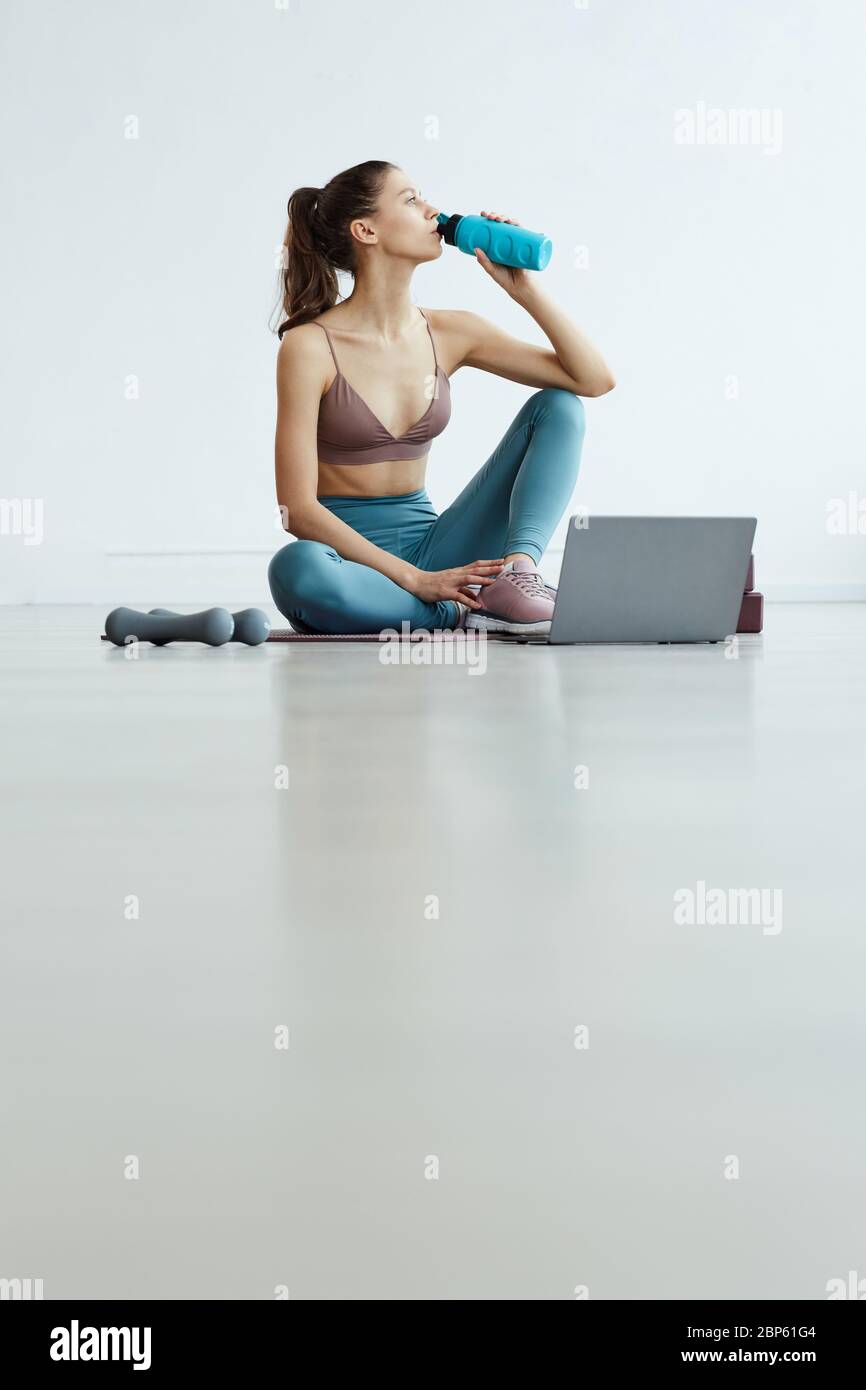 Ritratto verticale grandangolare di una giovane donna che beve acqua e guarda video sportivi tramite laptop durante l'allenamento a casa in interni bianchi minimal, COP Foto Stock