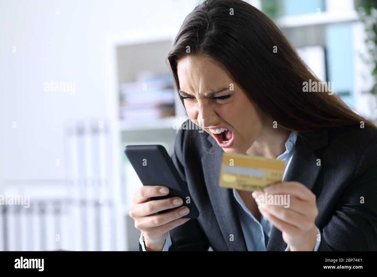 Donna esecutiva arrabbiata che gridava pagando online su smartphone con carta di credito seduta in ufficio Foto Stock