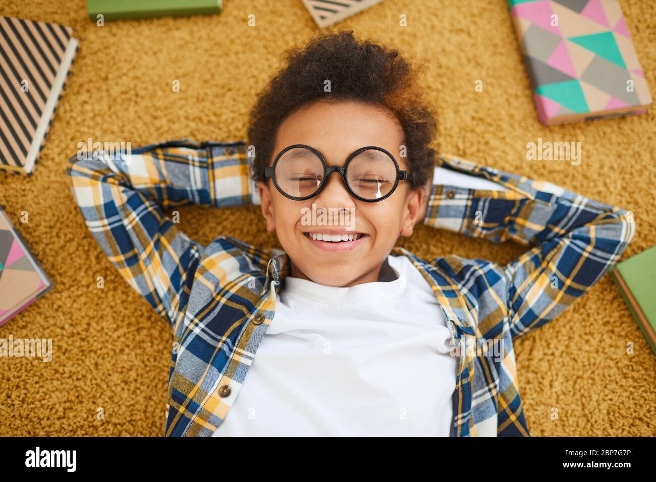 Sopra la vista ritratto di carino ragazzo africano indossare occhiali e sorridente alla macchina fotografica mentre si trova sul pavimento circondato da libri, spazio copia Foto Stock