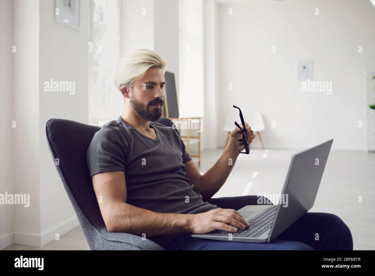 Lavorare online a casa. Formazione online. Un giovane uomo d'affari in abiti casual lavora con un computer portatile mentre si siede in una poltrona in una stanza residenziale. Foto Stock