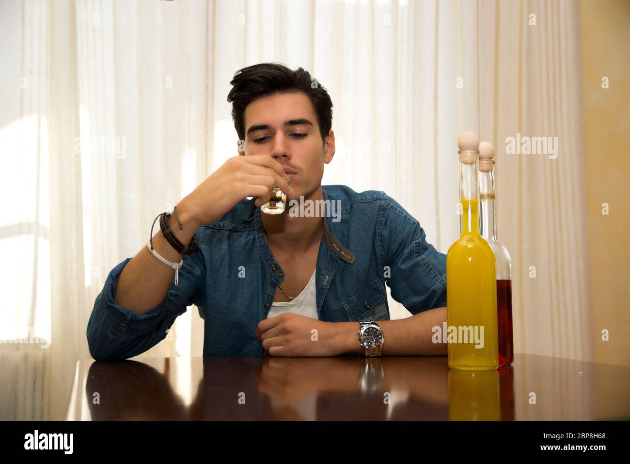 Giovane uomo seduto a bere da solo ad un tavolo con due bottiglie di liquori accanto lui sorseggiando dal colpo di vetro di affogare i suoi dispiaceri Foto Stock
