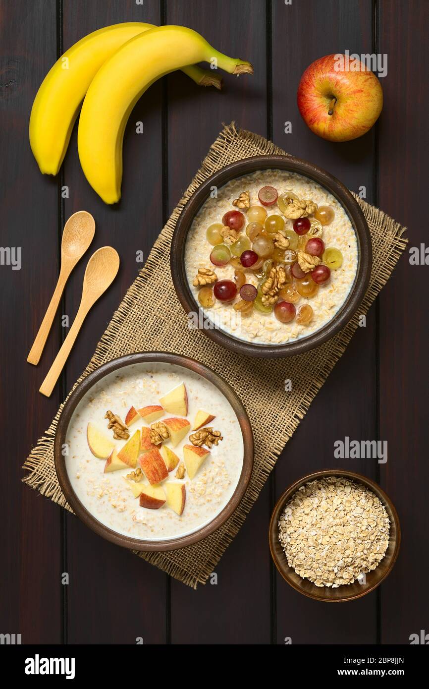 Porridge di farinata d'avena con uva, mele e noci in ciotole rustiche, mela fresca, banane, cucchiai di legno e farinata d'avena cruda sul lato, fotografato overhe Foto Stock