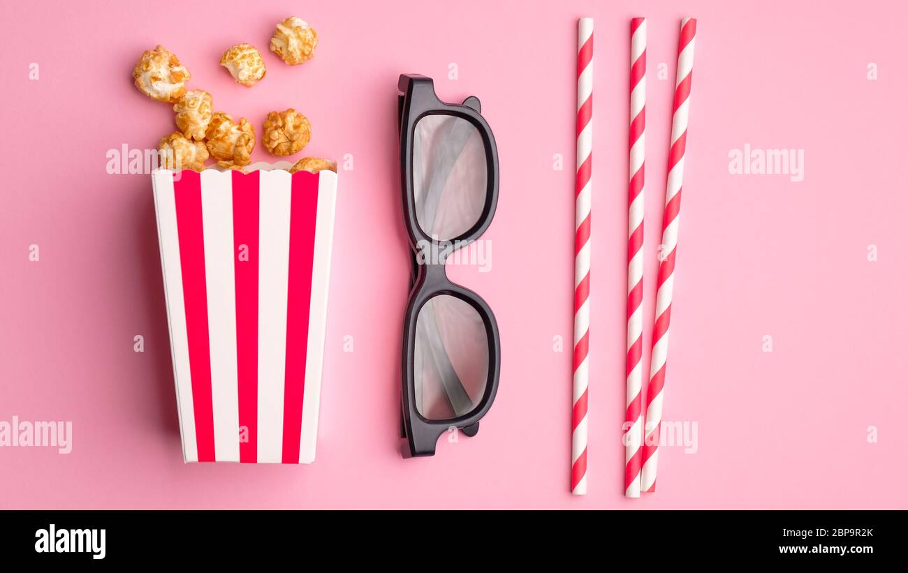 Composizione piatta con popcorn in scatola di cartone a righe, bicchieri 3d e cannucce da bibita su sfondo rosa. Visione di film al cinema. Foto Stock