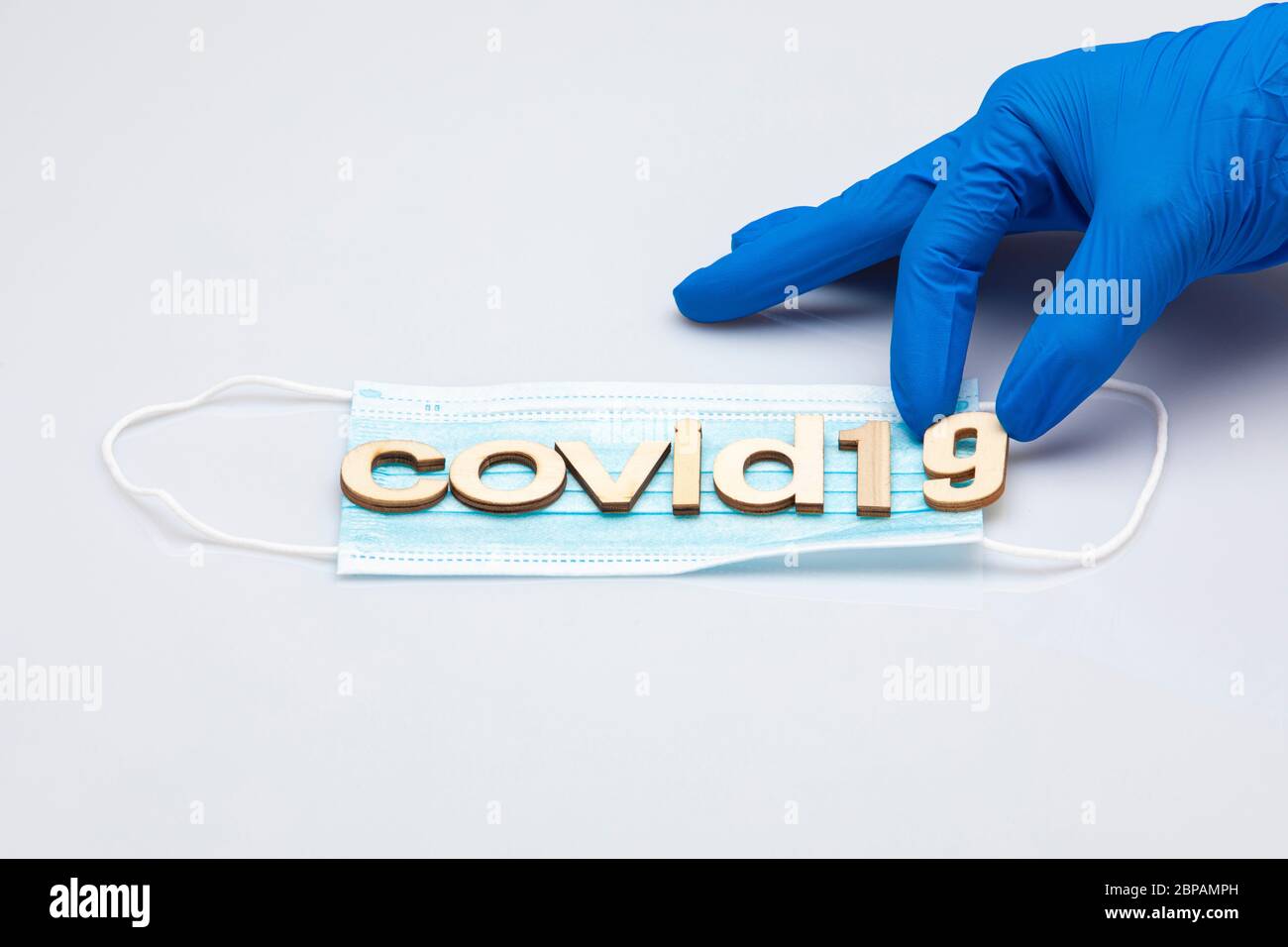 mano con un guanto chirurgico blu che tiene l'ultima parte della parola covid19 formato da lettere poste su una maschera chirurgica isolata su uno sfondo bianco Foto Stock