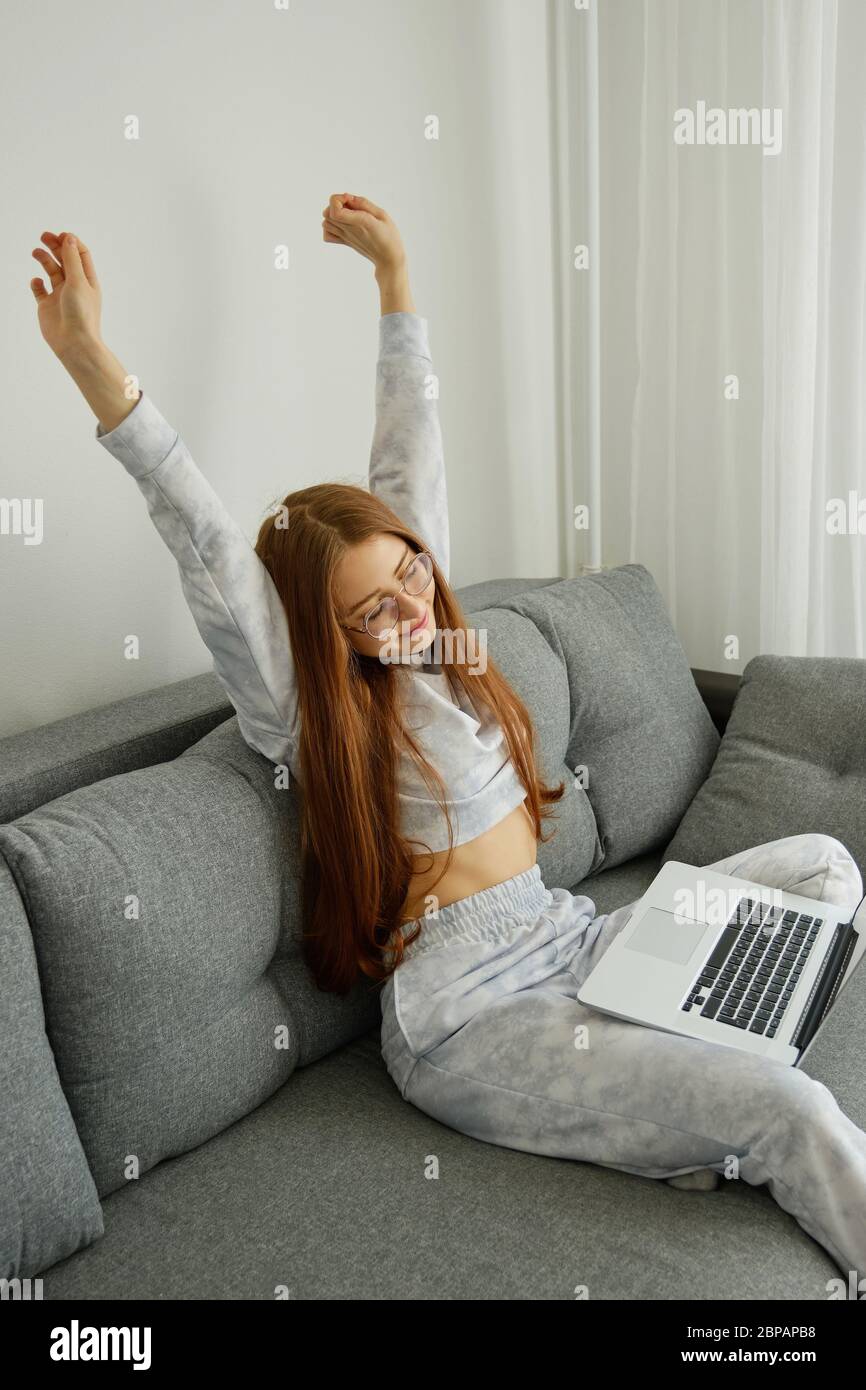 Ragazza rossa in pigiami e bicchieri si siede su un divano con un computer portatile, allungando le braccia Foto Stock