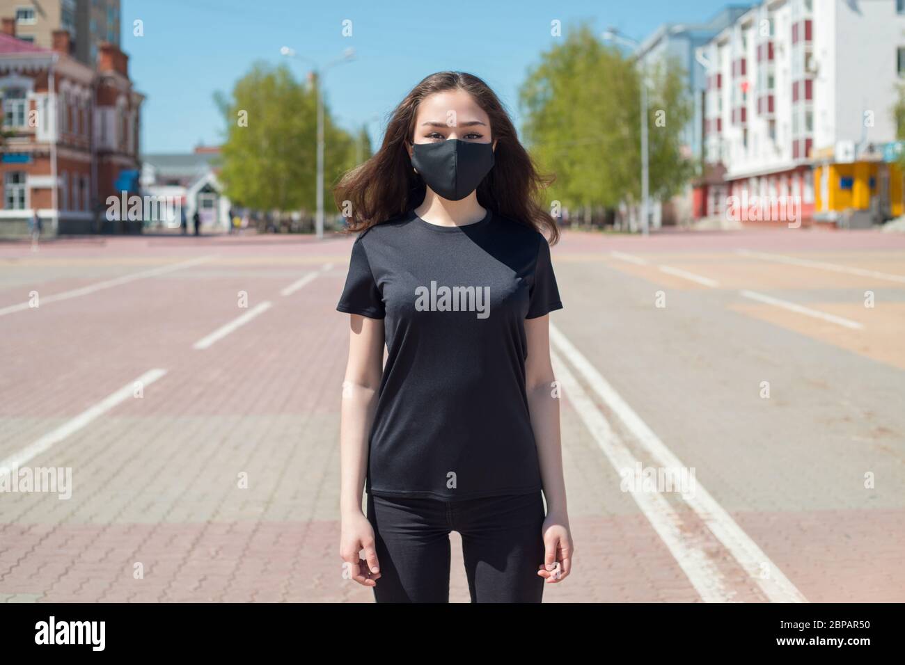 Giovane donna in una t-shirt nera e una maschera protettiva si trova sulla strada. Foto Stock