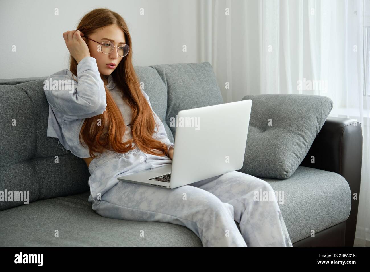 Ragazza rossa in pigiami e bicchieri si siede su un divano con un computer portatile, raddrizzando i capelli Foto Stock
