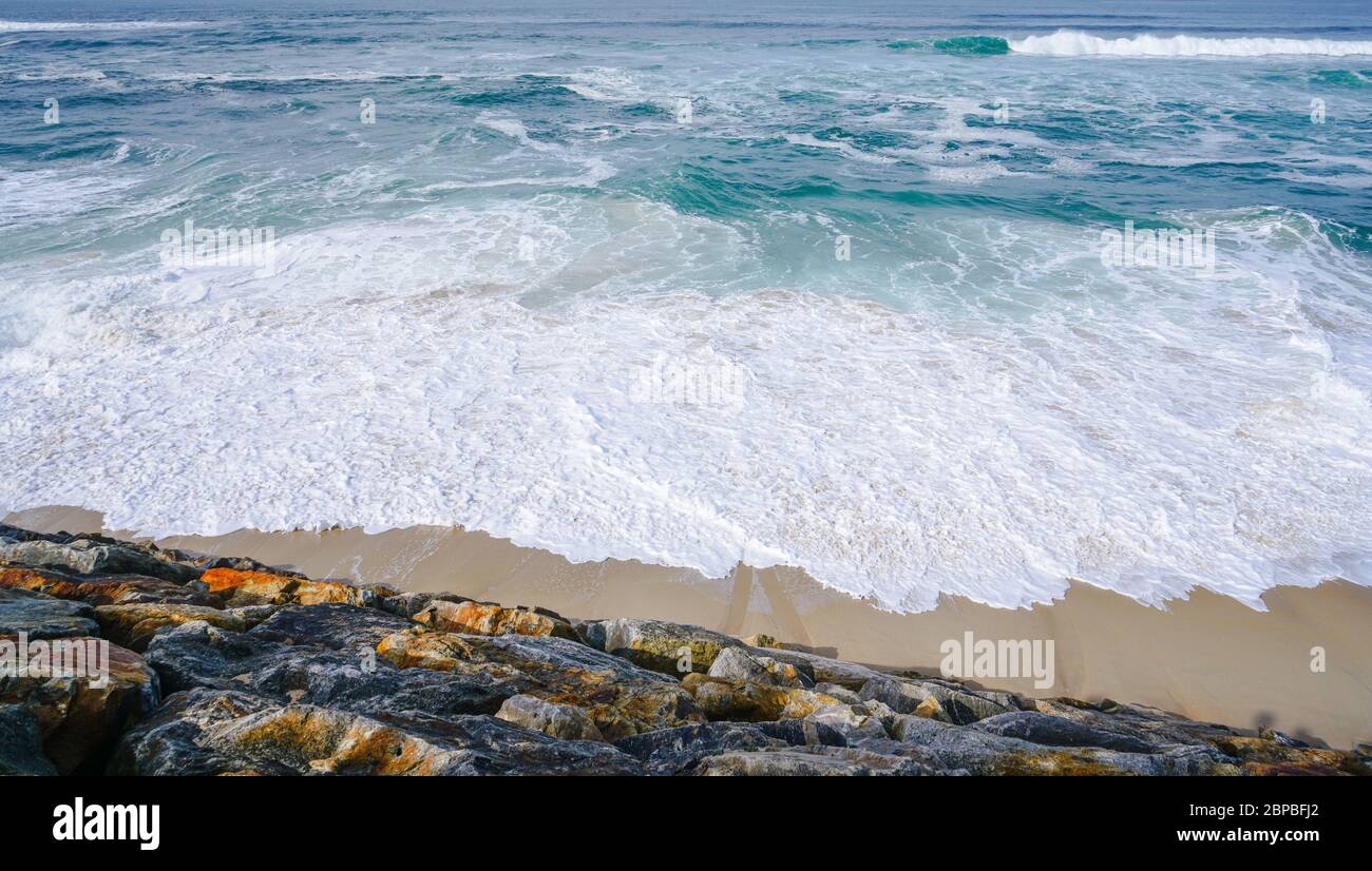 Acque turchesi dell'oceano con piccole onde bianche che si infrangono delicatamente sulla spiaggia di sabbia dorata e su una riva rocciosa Foto Stock