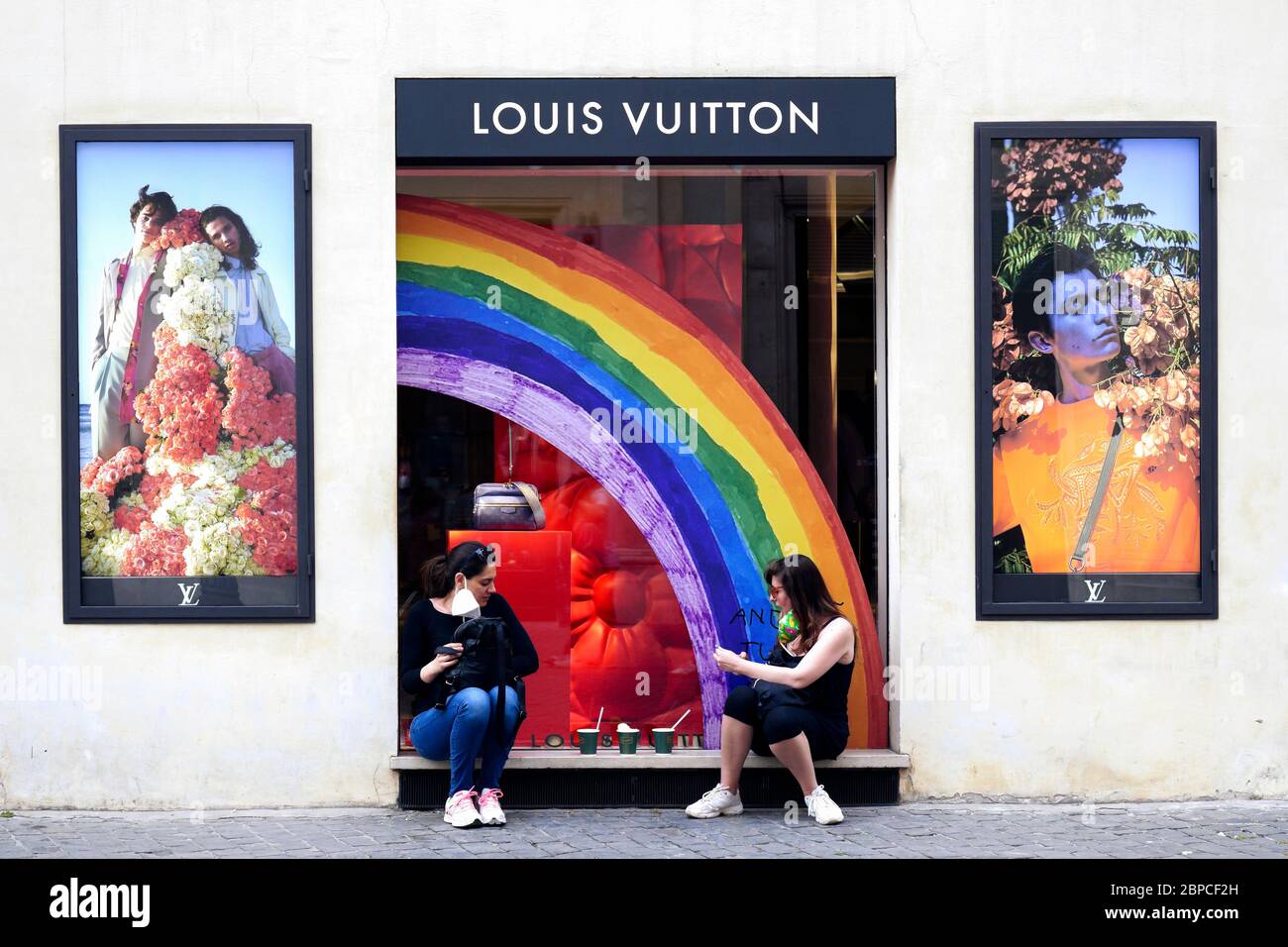 Due giovani amiche seduti di fronte alla finestra del negozio di bandiera Louis Vuitton, fare una pausa rilassante e togliere la loro maschera medica durante il blocco per coronavirus covid 19. Roma, Italia, Europa, Unione europea, UE. Foto Stock