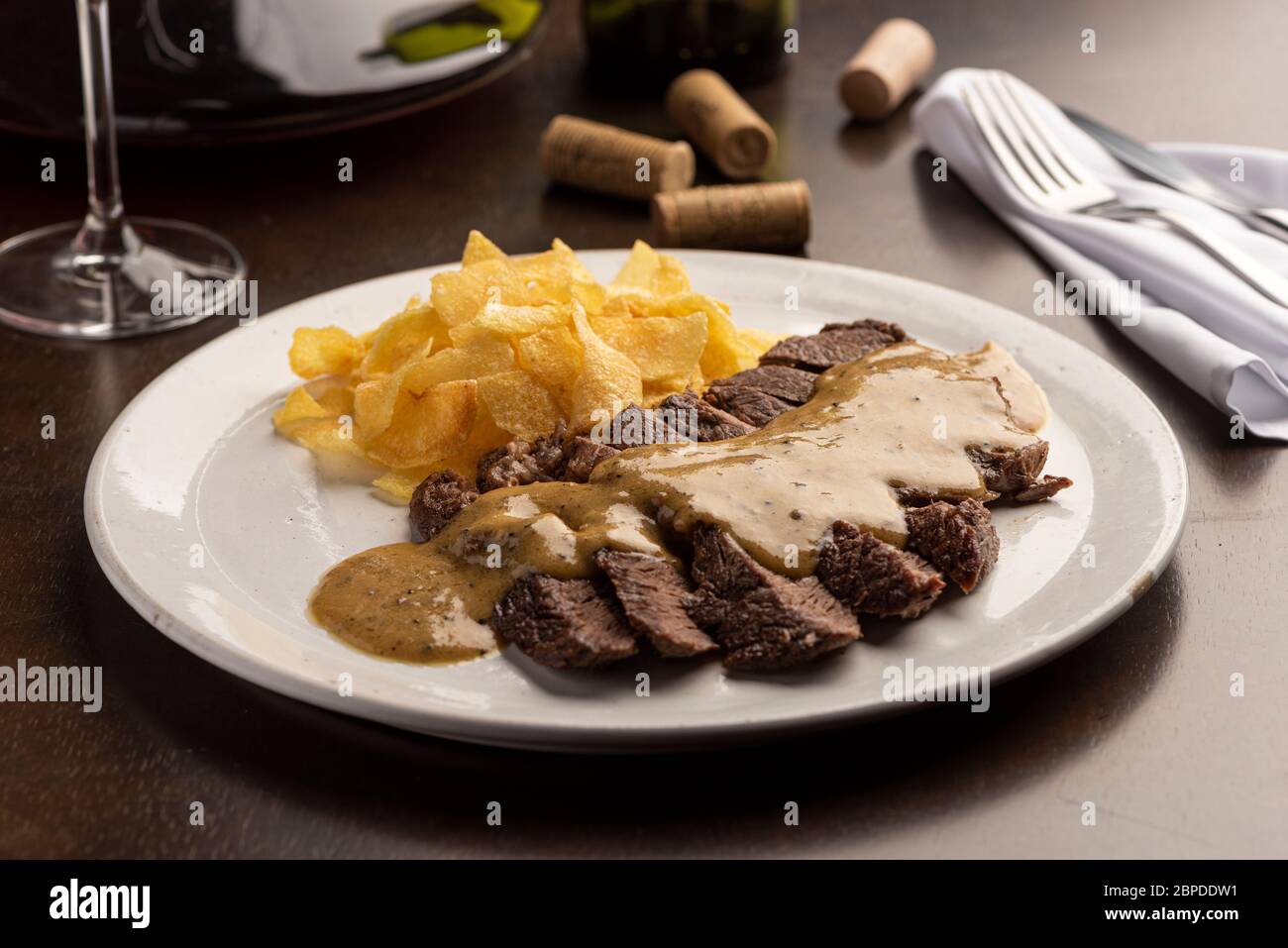 Bistecca con salsa con patatine su piatto bianco e bicchiere di vino, sughero, argenteria e tovagliolo Foto Stock