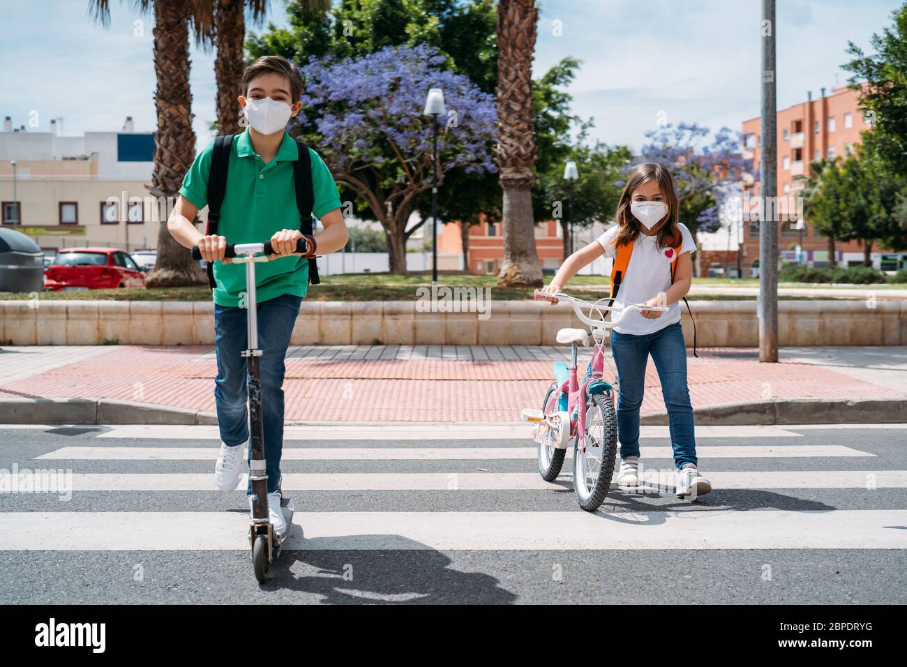 Ragazzo e ragazza che indossano maschere e cavalcano uno scooter e una bicicletta sulla strada Foto Stock