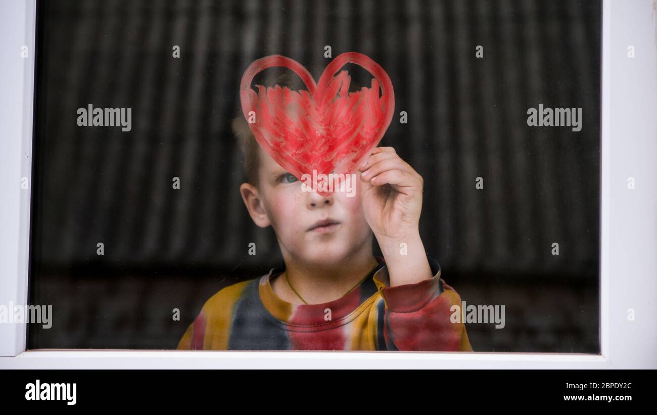 Ragazzo vicino alla finestra, arcobaleno, fine della quarantena. Un ragazzo disegna un arcobaleno e un cuore rosso. Il concetto di inseguimento dell'arcobaleno. Spazio copia per testo l Foto Stock