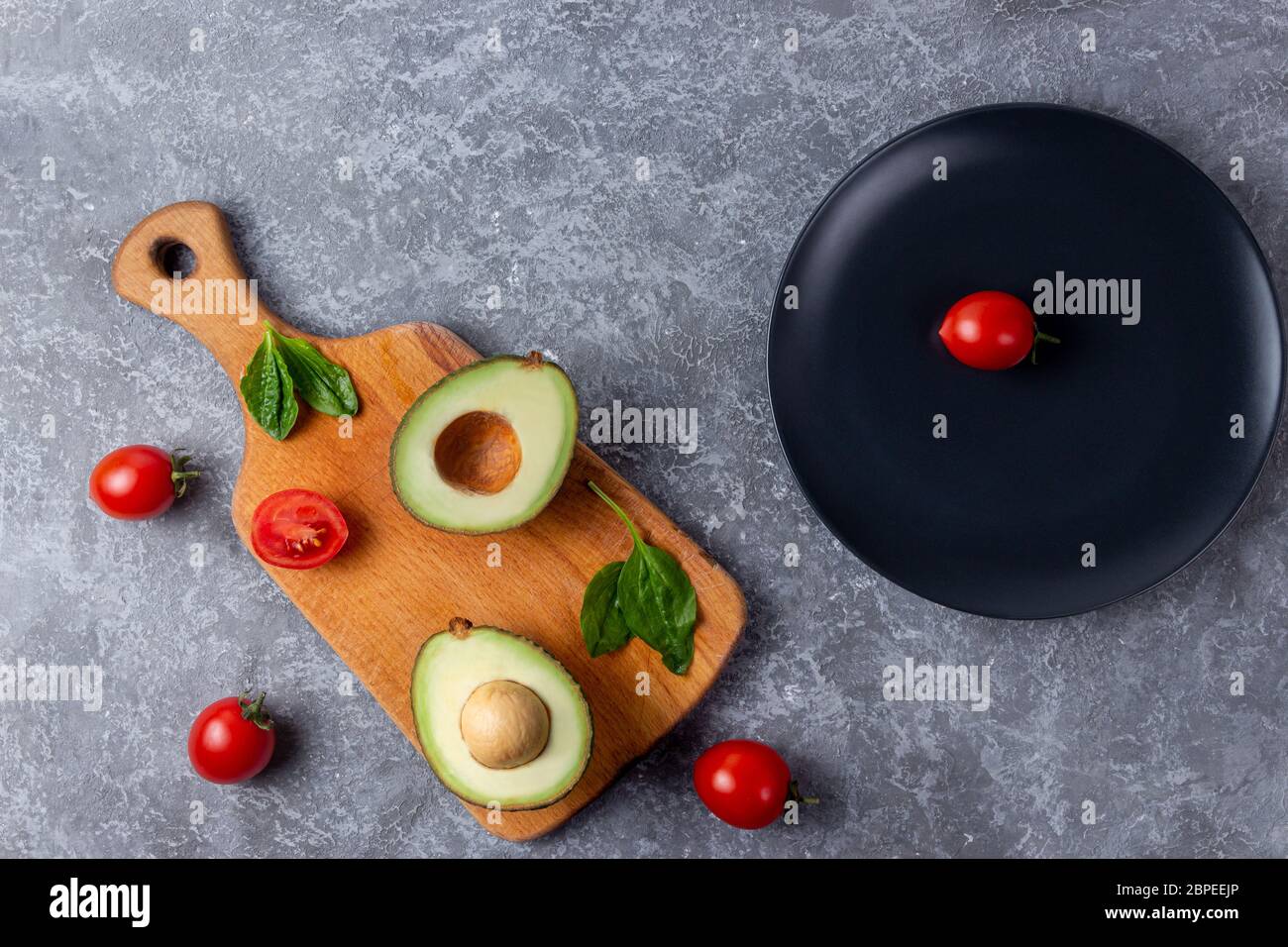 Avocado maturo tagliato per due parti con pomodoro di ciliegio su tavola di legno vicino al piatto nero con una ciliegia di pomodoro Foto Stock