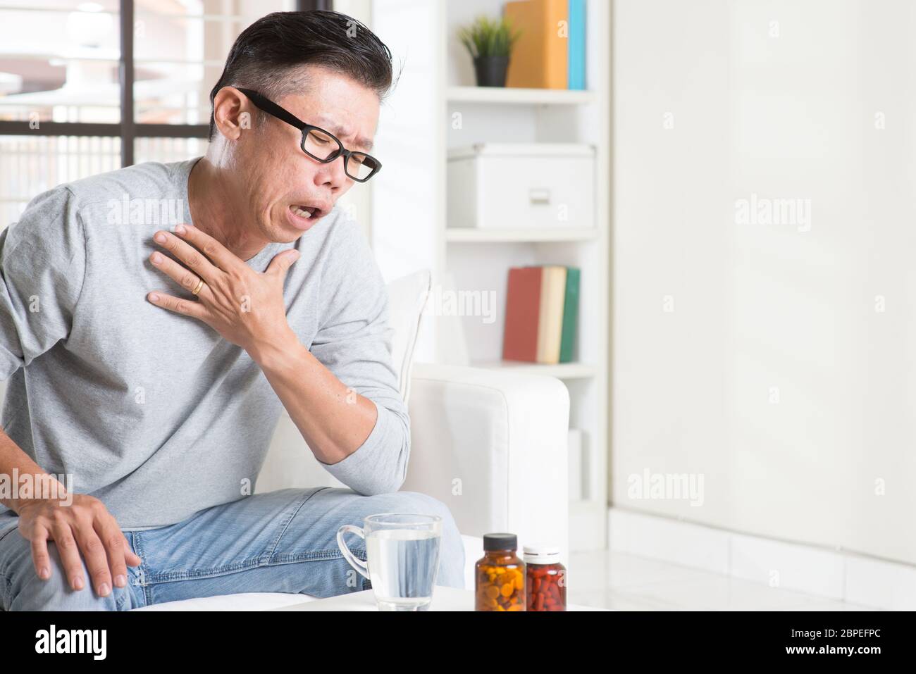Ritratto di casual 50s coppia uomo asiatico tosse, premendo sul petto con espressione dolorosa, seduto sul divano di casa, medicinali e acqua sul tavolo. Foto Stock
