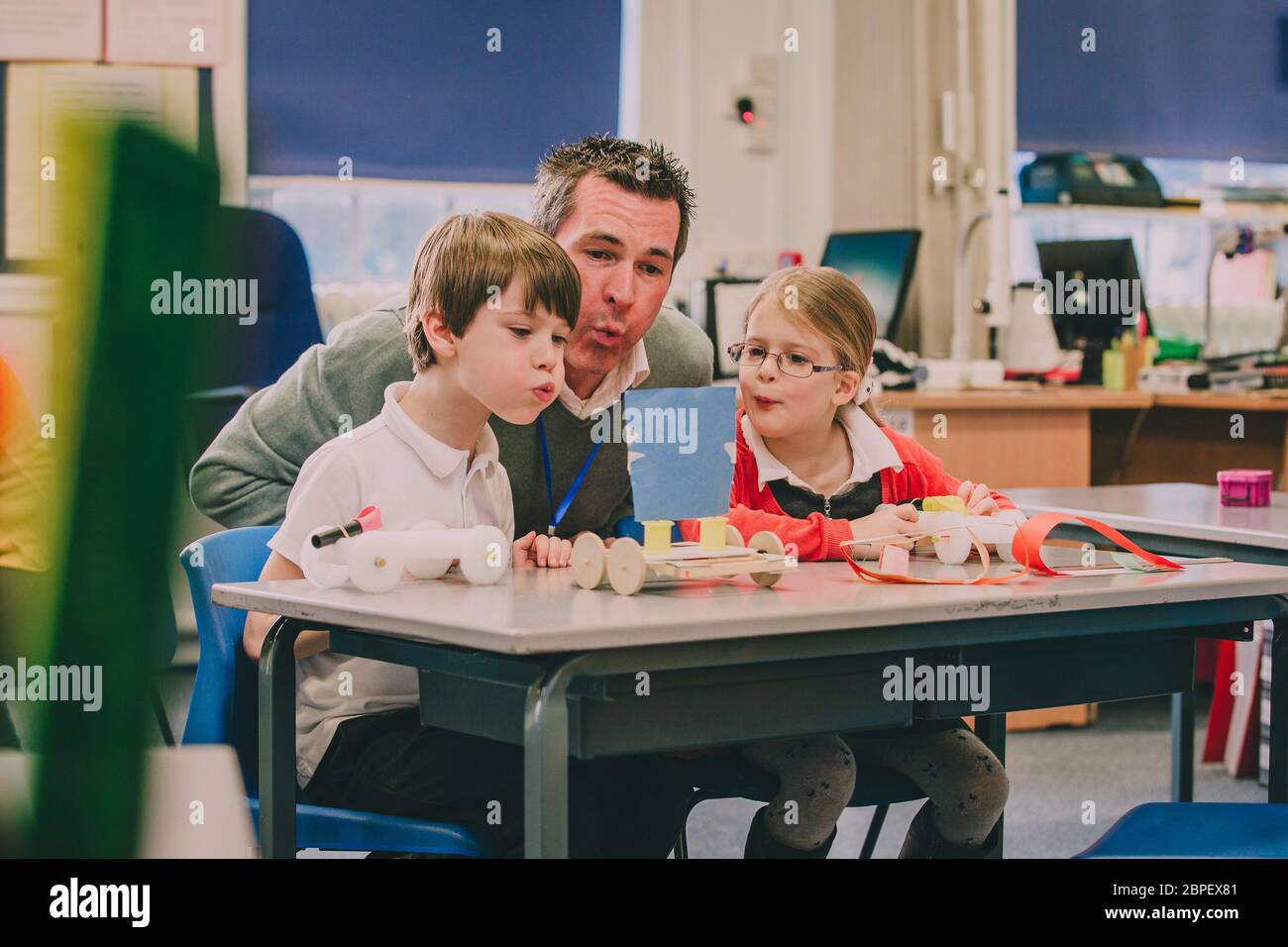 Insegnante è seduto con due dei suoi studenti della scuola primaria in aula, aiutandoli a prova il loro progetto di fisica. Essi sono la creazione di aria mediante soffiaggio Foto Stock