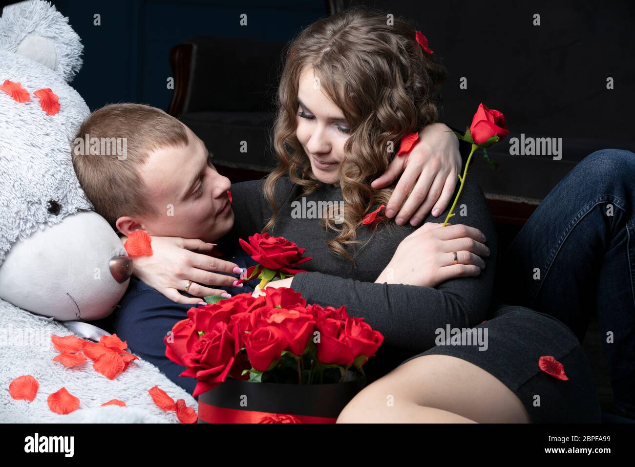 Bella giovane coppia a casa. Abbracci, bacia e ama trascorrere il tempo insieme, festeggiando San Valentino con rose rosse su un orsacchiotto. Foto Stock