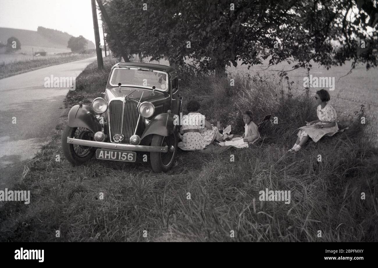 In una zona erbosa, accanto a una strada di campagna vuota, una madre in un vestito macchiato siede dal lato passeggero di una vettura, un salone sportivo dell'epoca che ha un picnic con le sue due ragazze entrambe nei loro abiti estivi, circa 1940, Inghilterra, UK. Foto Stock
