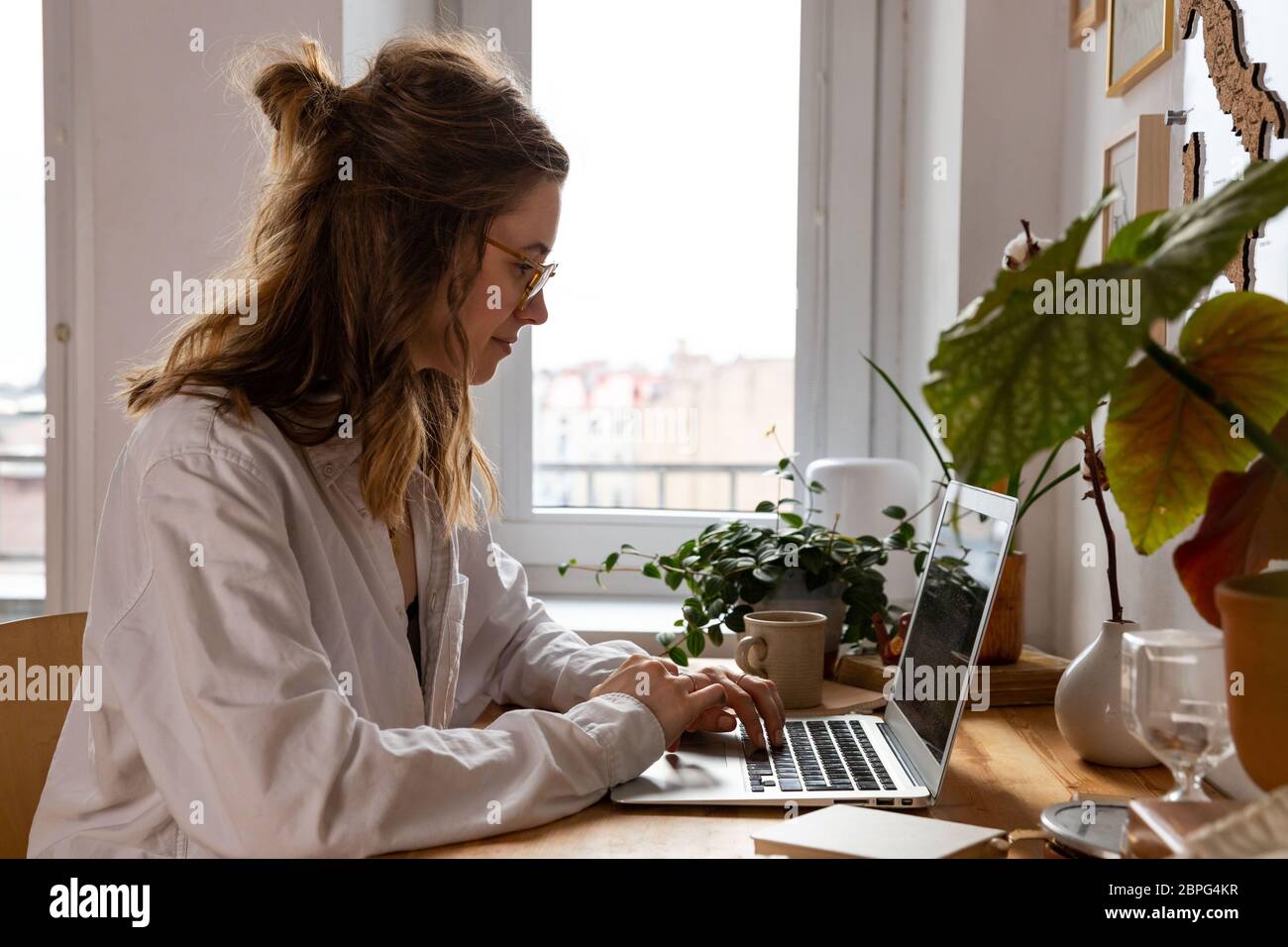 Giovane donna freelance/designer che lavora al computer da casa durante l'auto-isolamento a causa del coronavirus. Ambiente di lavoro accogliente circondato da piante. Re Foto Stock