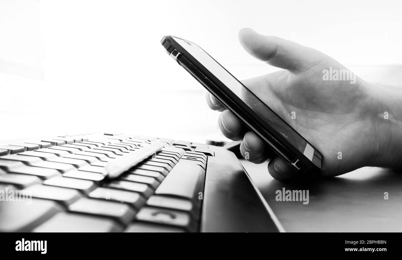 La mano dell'uomo tiene uno smartphone su una tastiera nera del computer. Sfondo molto sfocato. Immagine in bianco e nero. Spazio di copia. Foto Stock