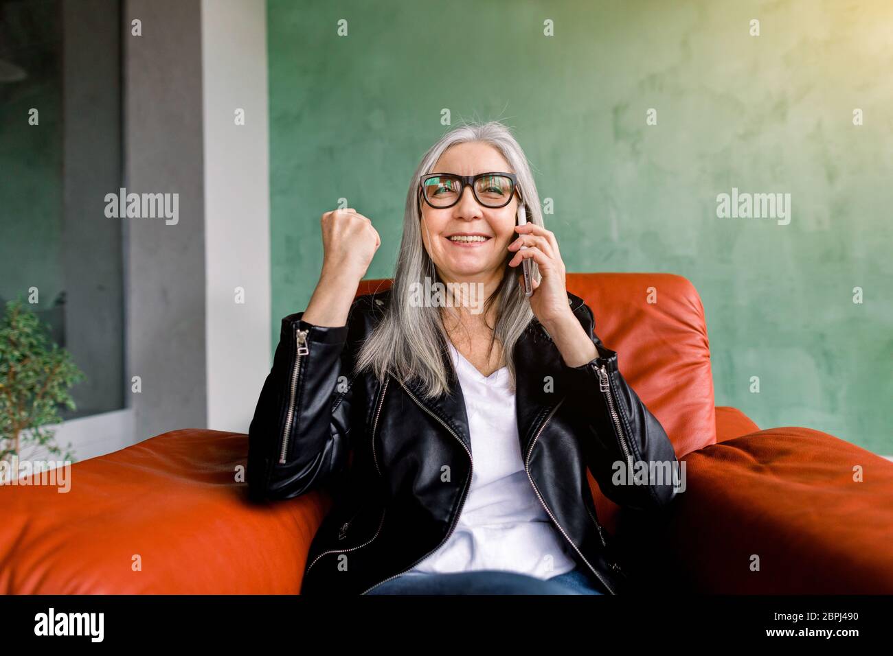 Bella donna anziana moderna in giacca nera in pelle, seduta in morbida  poltrona rossa, mantenendo la sua pugno clenched e sorridendo durante la  chiamata mobile con lei Foto stock - Alamy