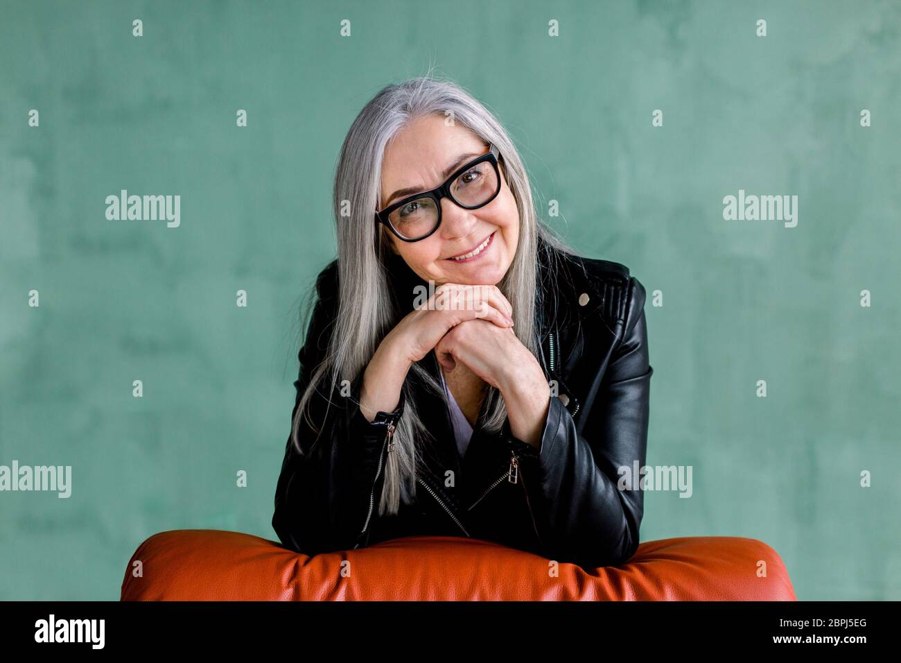 Studio ritratto di affascinante donna moderna anziana in occhiali, con lunghi capelli grigi dritti, indossando giacca trendy in pelle nera, che posa su Foto Stock