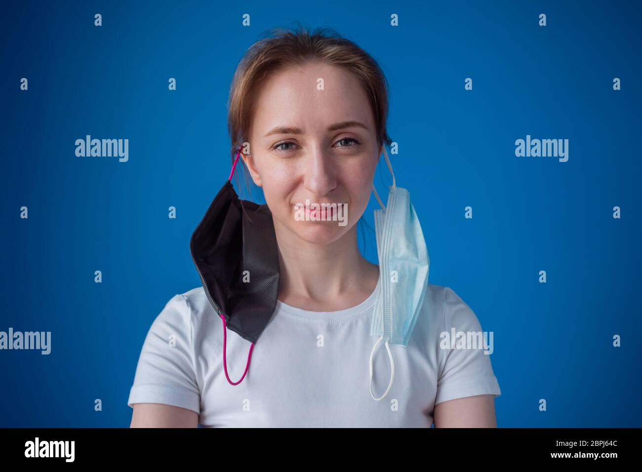 Ritratto di donna divertente con due maschera medica che pende sulle orecchie, guardando la macchina fotografica in camera con parete blu a casa. Autoisolamento, prevenzione Foto Stock