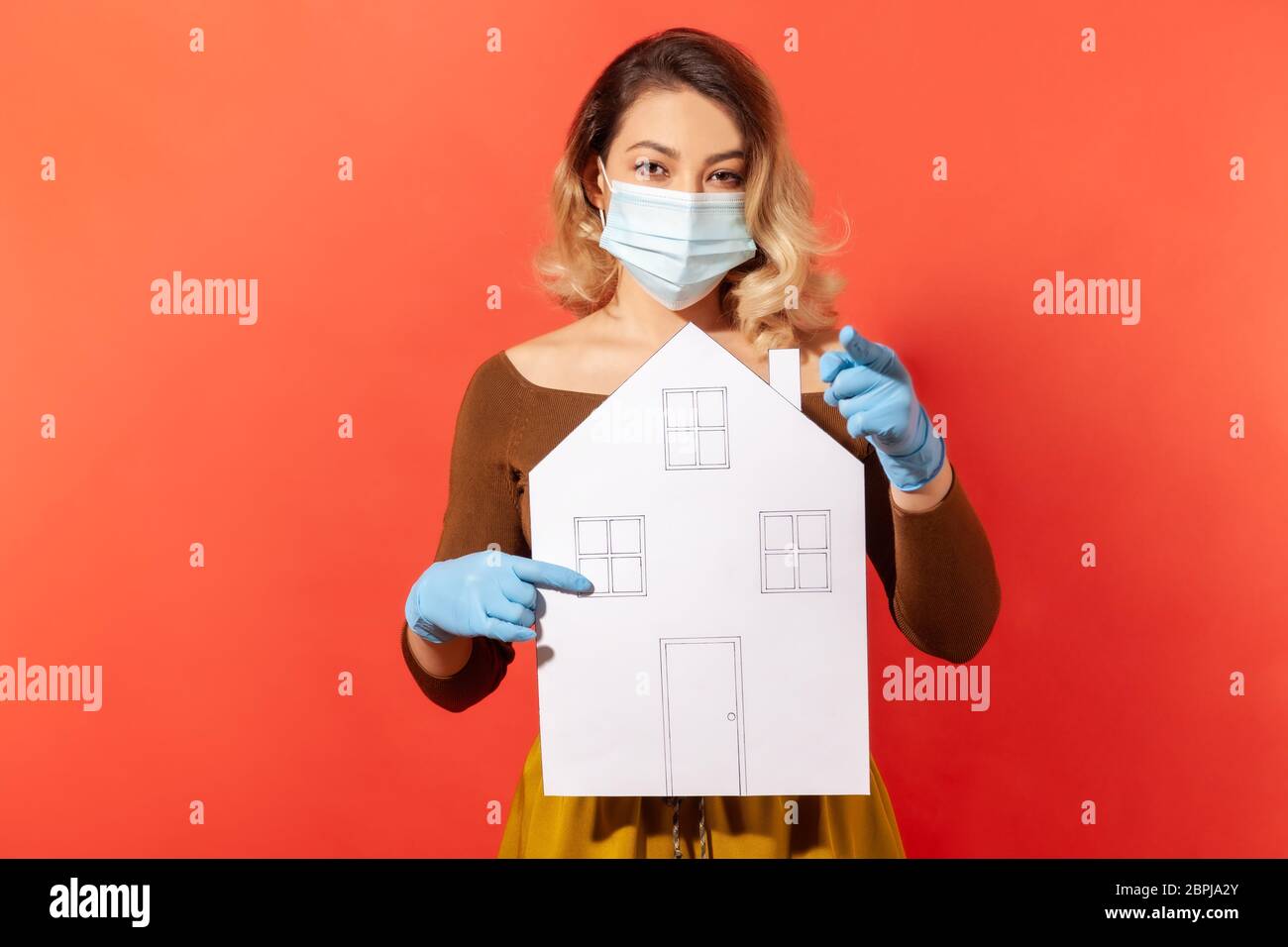 Rimani a casa! Donna in maschera chirurgica e guanti che tengono la casa di carta, puntando il dito alla macchina fotografica, raccomandando auto-isolamento, quarantena mentre co Foto Stock
