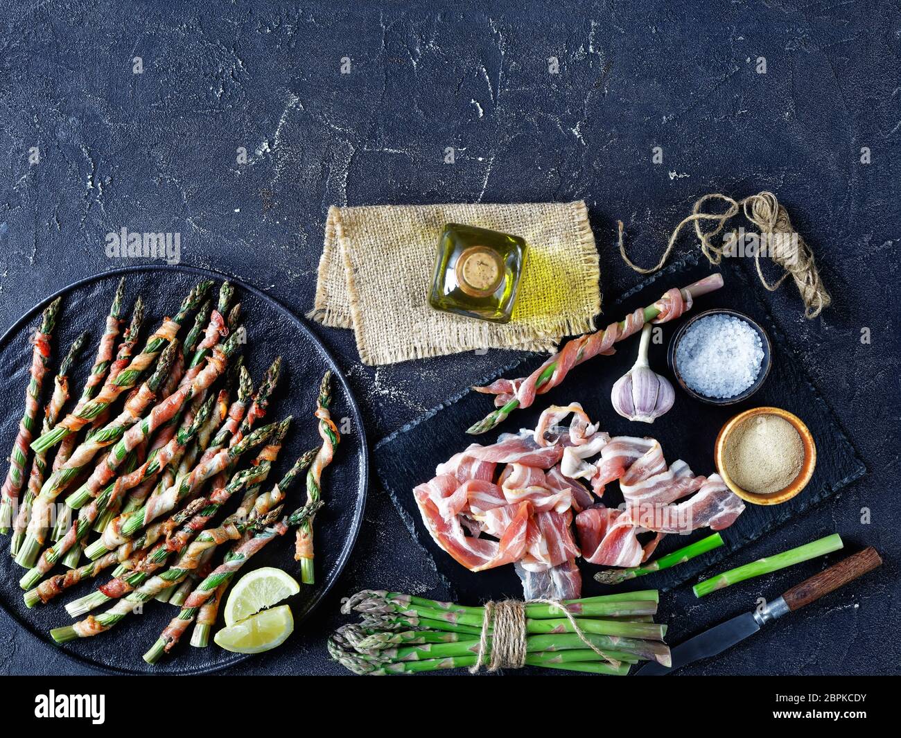 Asparagi avvolti con pancetta cotti al forno con aglio e sale marino serviti su un piatto nero con olio d'oliva su fondo di cemento scuro, vista dall'alto, Fla Foto Stock