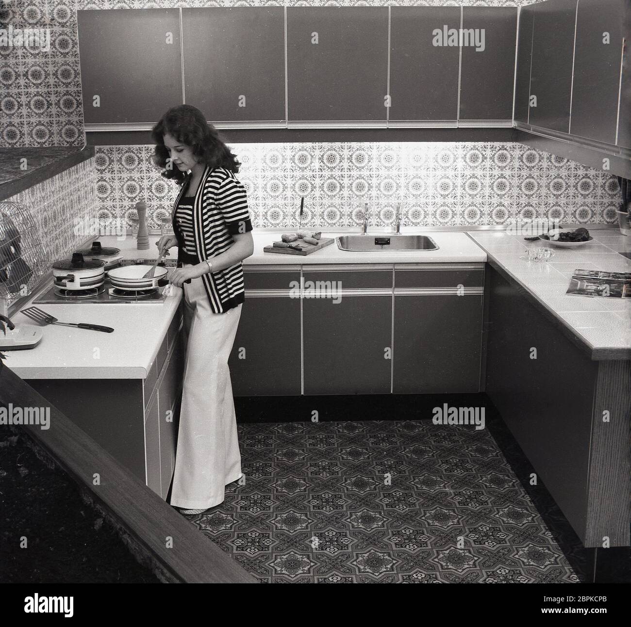 Anni '70, una donna elegante e di stile in un top stripy e pantaloni svasati che cucina in una cucina compatta e moderna dell'epoca, con unità da cucina in laminato e countertops, splashbacks e pavimento a motivi geometrici, Inghilterra, Regno Unito. Foto Stock