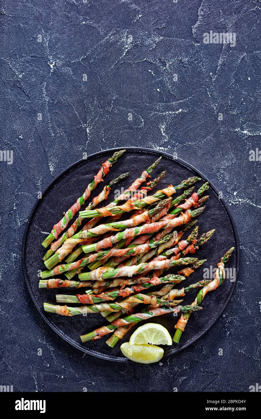 Verdure primaverili: Asparagi avvolti in pancetta e grigliati con aglio e sale marino serviti su un piatto nero con olio d'oliva, limone su un bac di cemento scuro Foto Stock