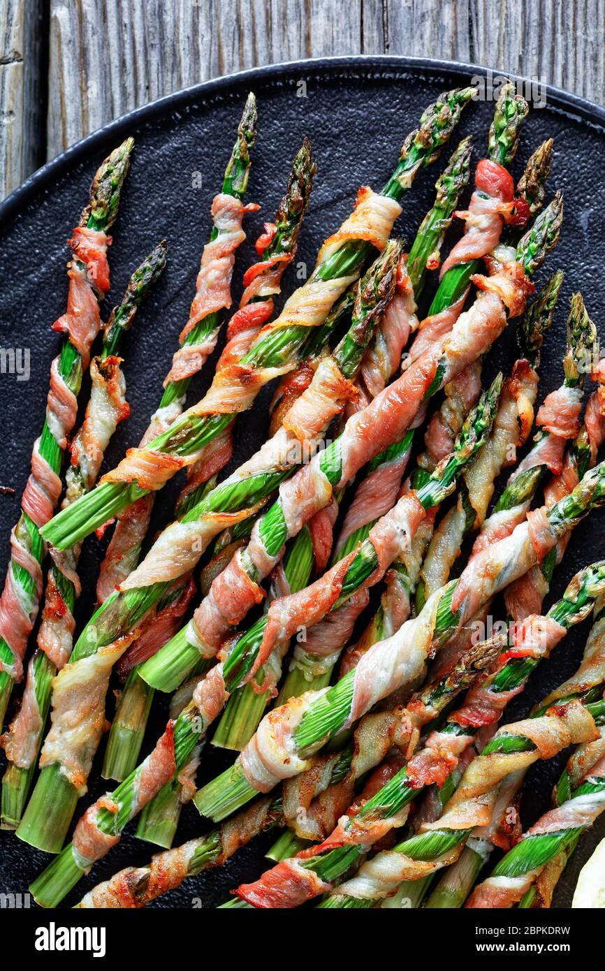 Piatto basso di carb pancetta avvolta asparagi facile contorno grigliato con aglio e sale marino servito su un piatto nero con limone su un vecchio fondo di legno, Foto Stock