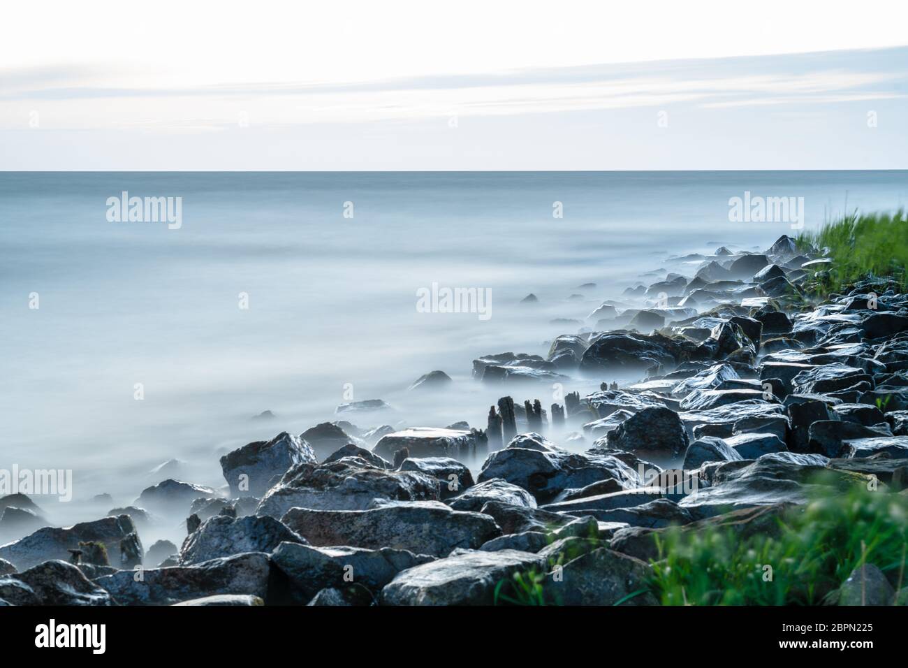 Mare costa con onde che si schiantano sulle rocce. Esposizione lunga, immagine sfocata dell'acqua, nebulosa tra le rocce sulla riva. Il cielo grigio sovrastato. Foto Stock