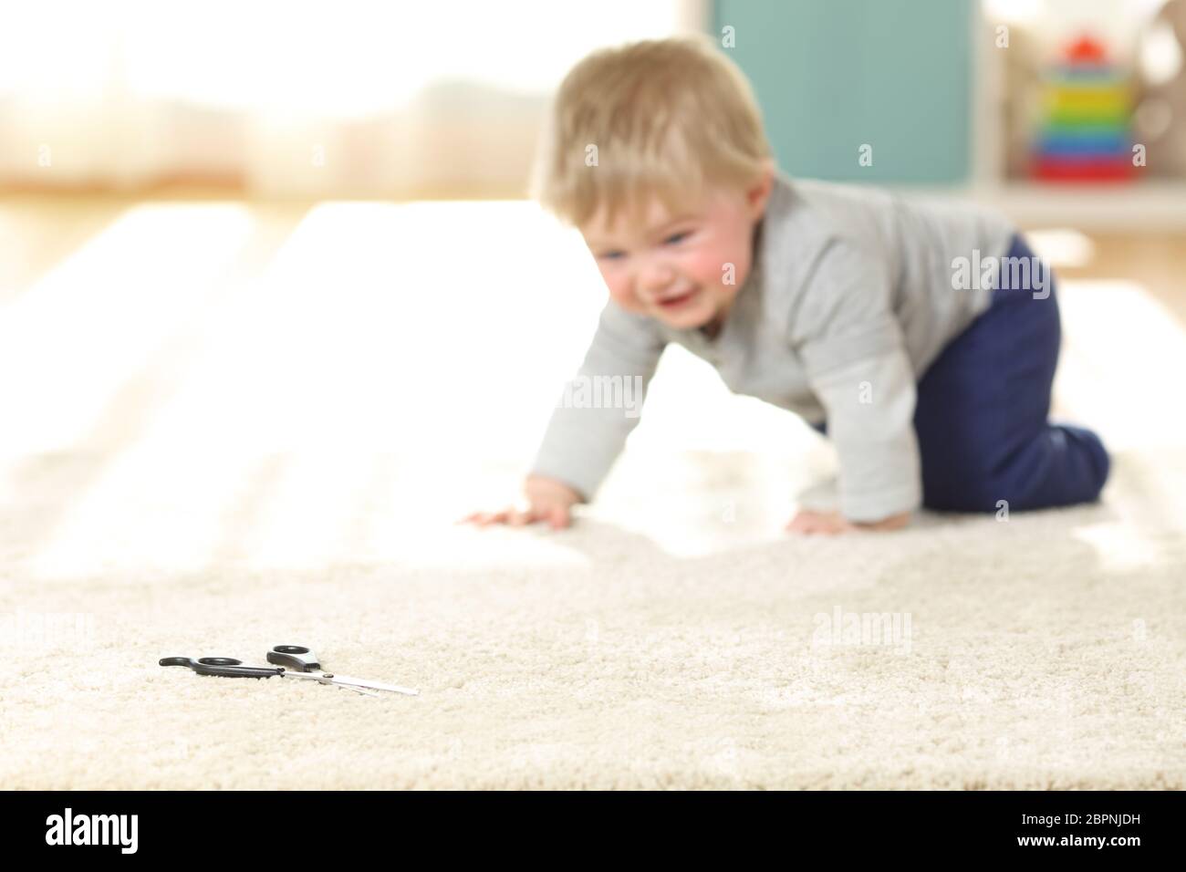 Bambino in pericolo strisciando verso una forbice su un tappeto a casa Foto Stock