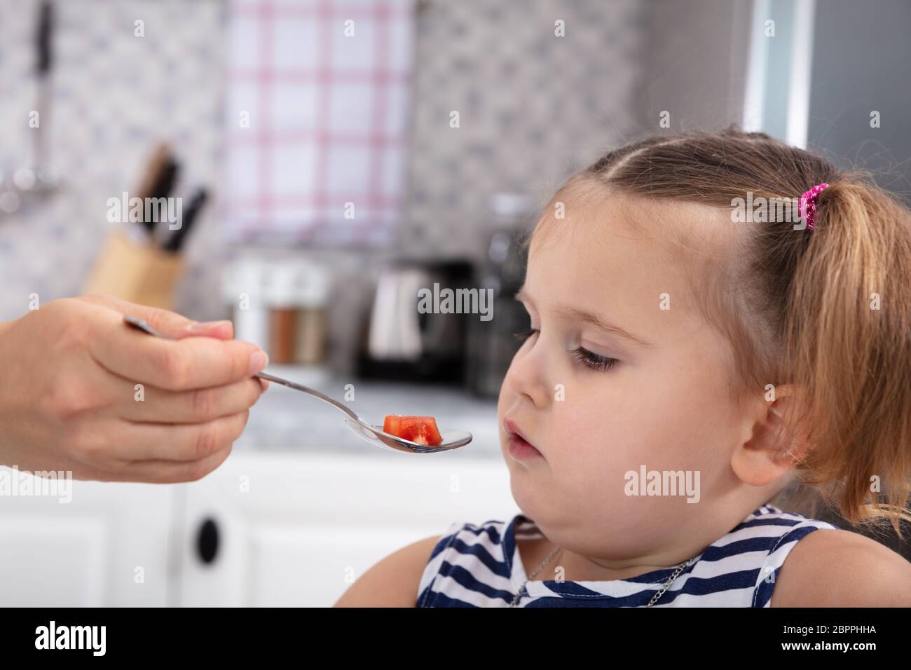 Close-up di Madre di alimentazione manuale fetta di pomodoro a sua figlia con cucchiaio in cucina Foto Stock