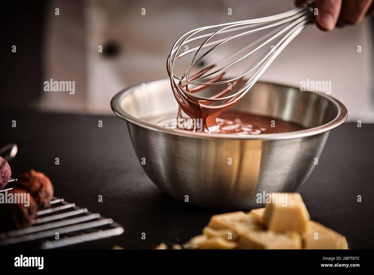 Lo Chef tenendo una frusta con il cioccolato fuso in un recipiente di miscelazione in cucina con ingredienti e bomboni a fianco Foto Stock