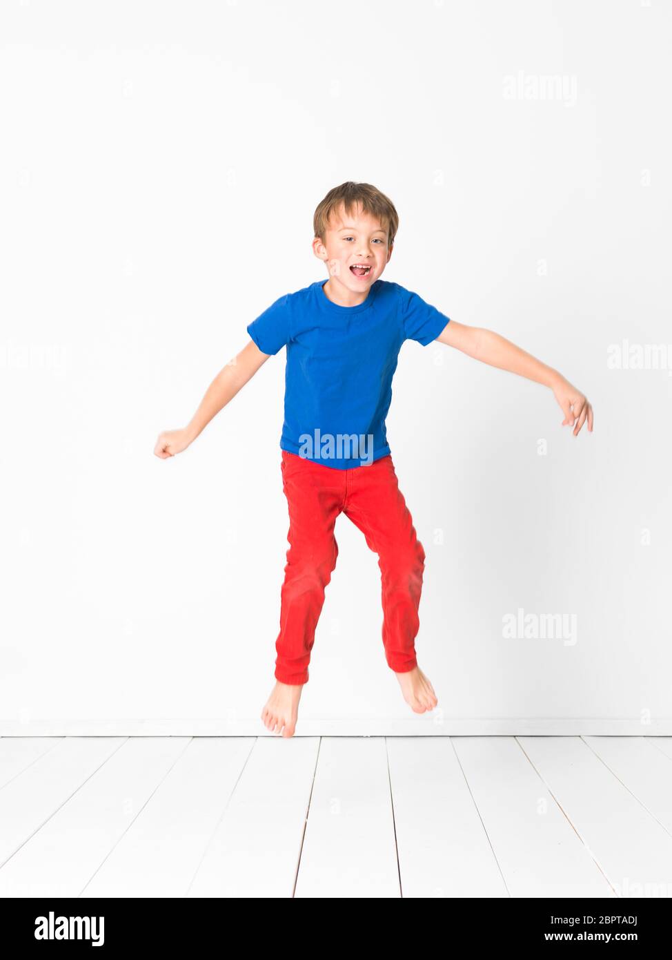 Cool, carino ragazzo con la maglietta blu e pantaloni rossi è il salto ad alta nello studio di fronte a uno sfondo bianco e bianco con pavimento in legno Foto Stock