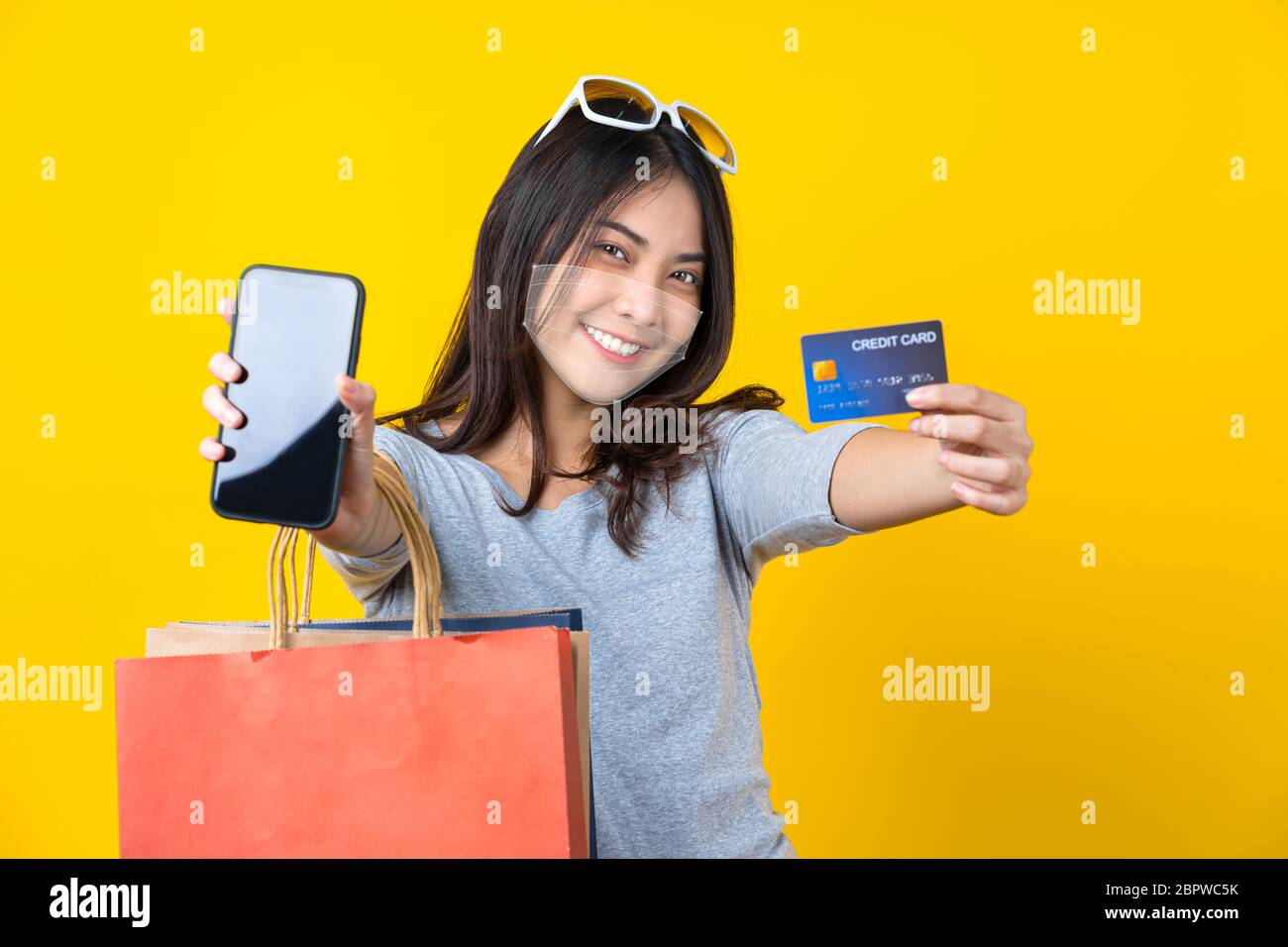 Happy Asian sorridente donna indossando tecnologia aumentata realtà maschera chirurgica in covid19 scoppio e presentando carta di credito e telefono cellulare con auto Foto Stock