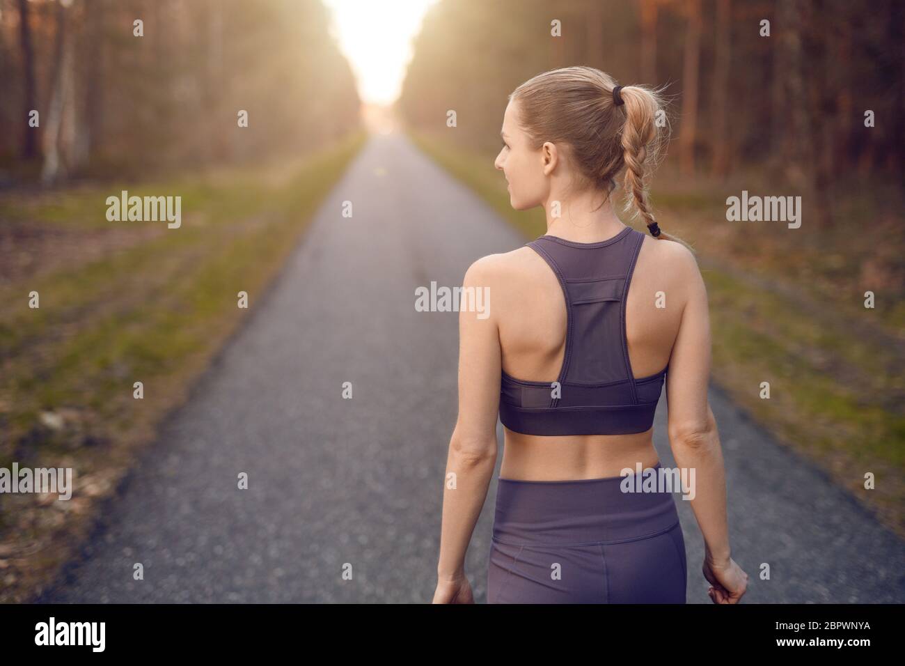 Giovane donna sportiva e sana che cammina all'alba lungo una strada rurale attraverso una fitta foresta verso il bagliore del sole alla fine tra gli alberi in un Foto Stock