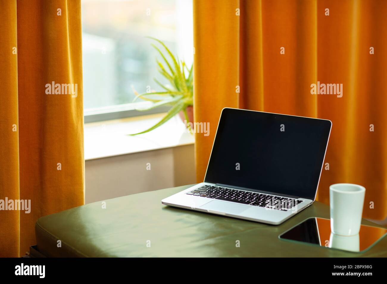 Un computer portatile che si stesa su un divano verde e una tenda arancione. Interni retrò dai colori caldi e una tazza di tè sul lato. Tecnologia moderna e. Foto Stock