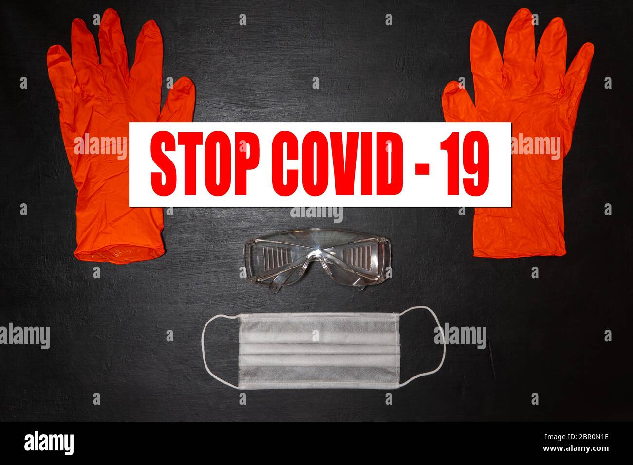 Sfondo medico nero. Occhiali di protezione, maschera medica e due guanti rossi. Sui guanti c'è un banner con le parole Stop Covid-19 Foto Stock