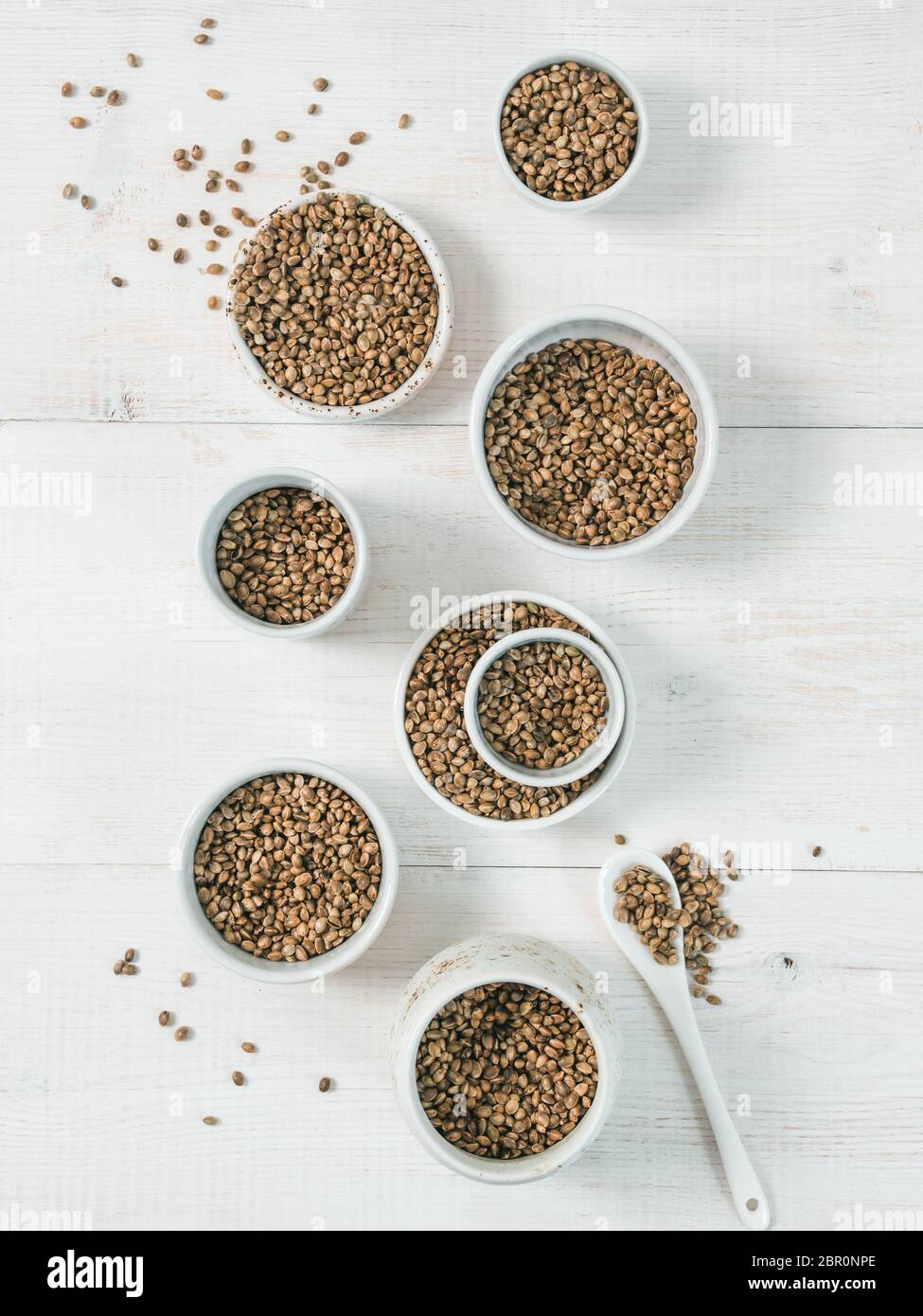 Sementi di canapa in piccole ciotole di bianco e cucchiaio bianco su un tavolo di legno. Set di piccole ciotole con organici grezzo grezzo di semi di canapa. Super e vegane concetto. Foto Stock